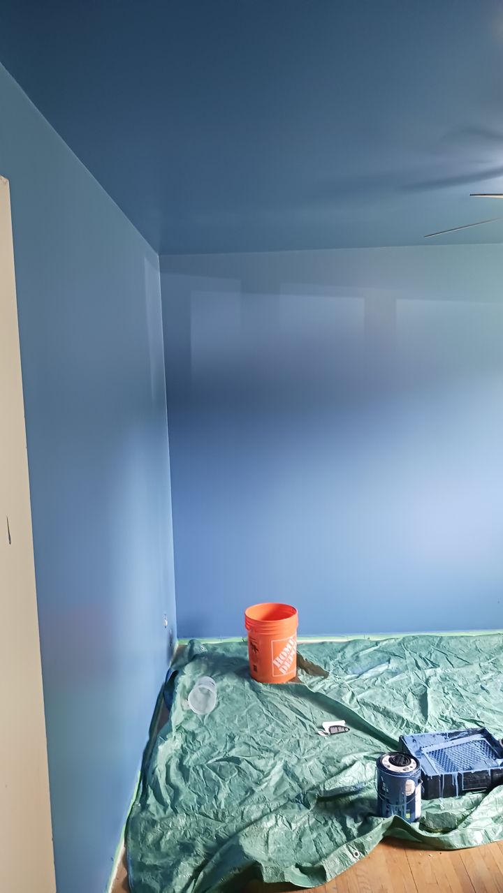 A room mid-paint: blue walls and ceiling, drop cloth and paint tray on the floor